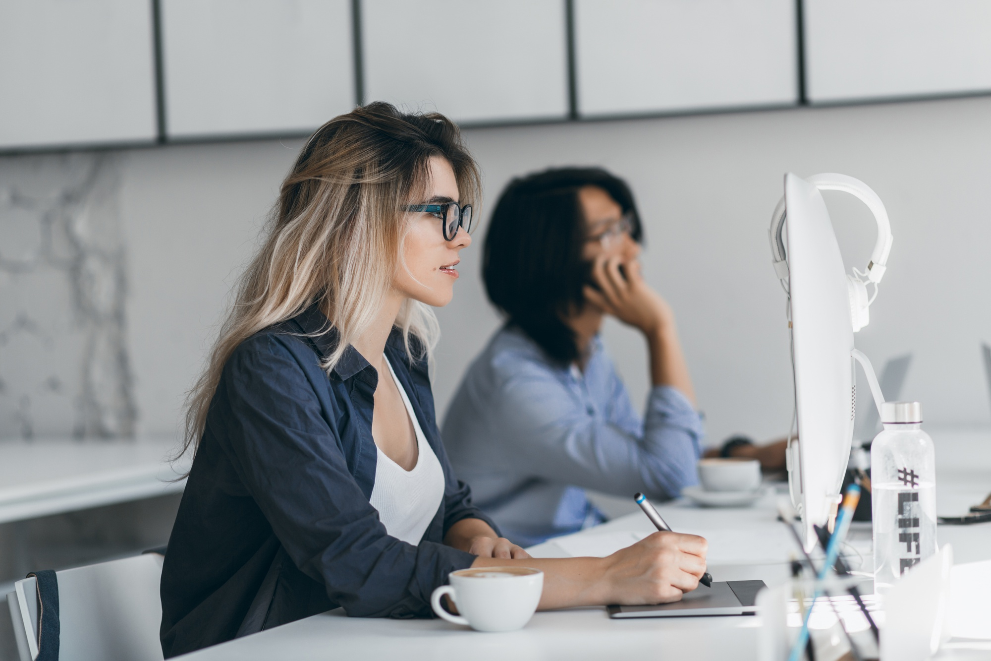 inspired-freelance-web-designer-using-tablet-stylus-looking-screen-while-her-friend-talking-phone-asian-student-holding-smartphone-typing-keyboard-sitting-beside-blonde-girl